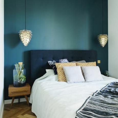 Stylowe wnętrze sypialni z małym drewnianym stolikiem nocnym, ogrodem w słoiku, białą pościelą, kolorowymi stosami i kocem. Przestrzeń z niebieskimi ścianami i brązowym drewnianym parkietem. Designerska lampa.