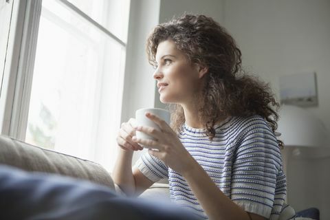 Uśmiechnięta młoda kobieta patrzeje z okno z filiżanką kawy