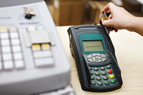 Ręka kobiety przesuwa kartę przez maszynę do kart kredytowych stojącą obok kasy. Nacisk kładziony jest na urządzenie do kart kredytowych.